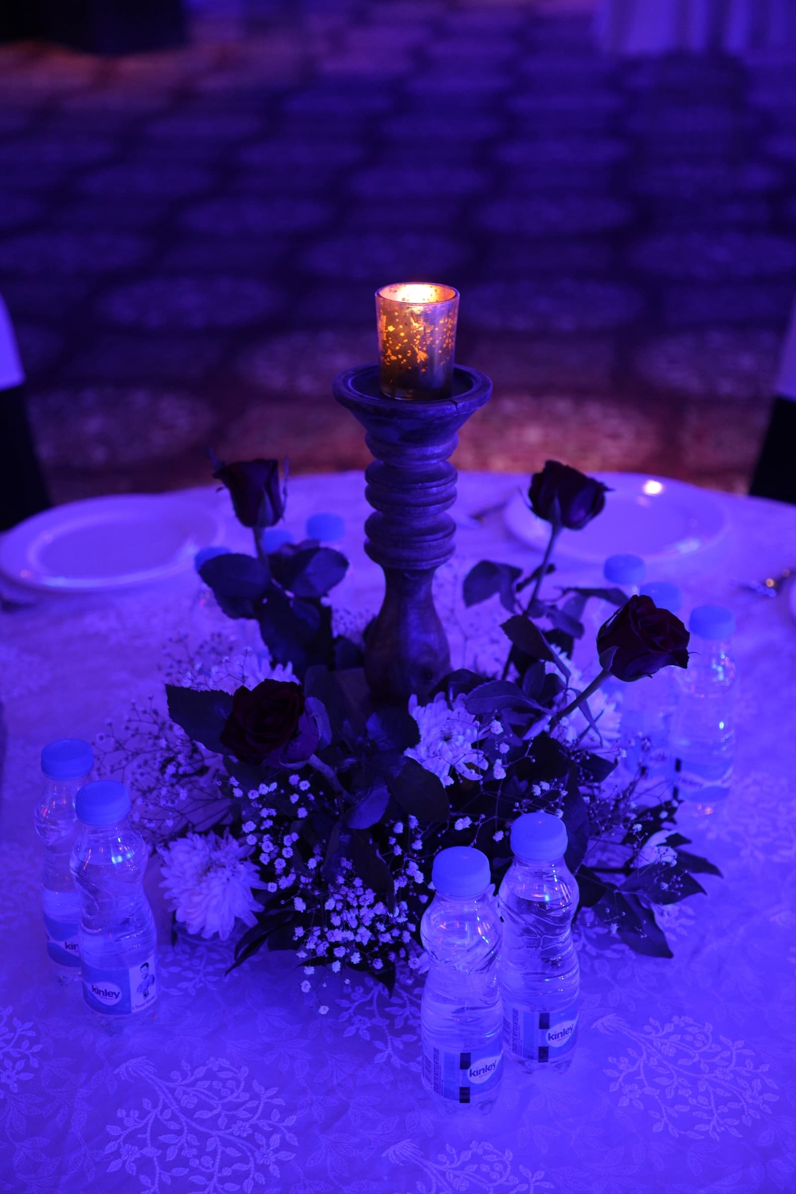 A candlelit table centrepiece of roses and baby's breath under blue cocktail lighting