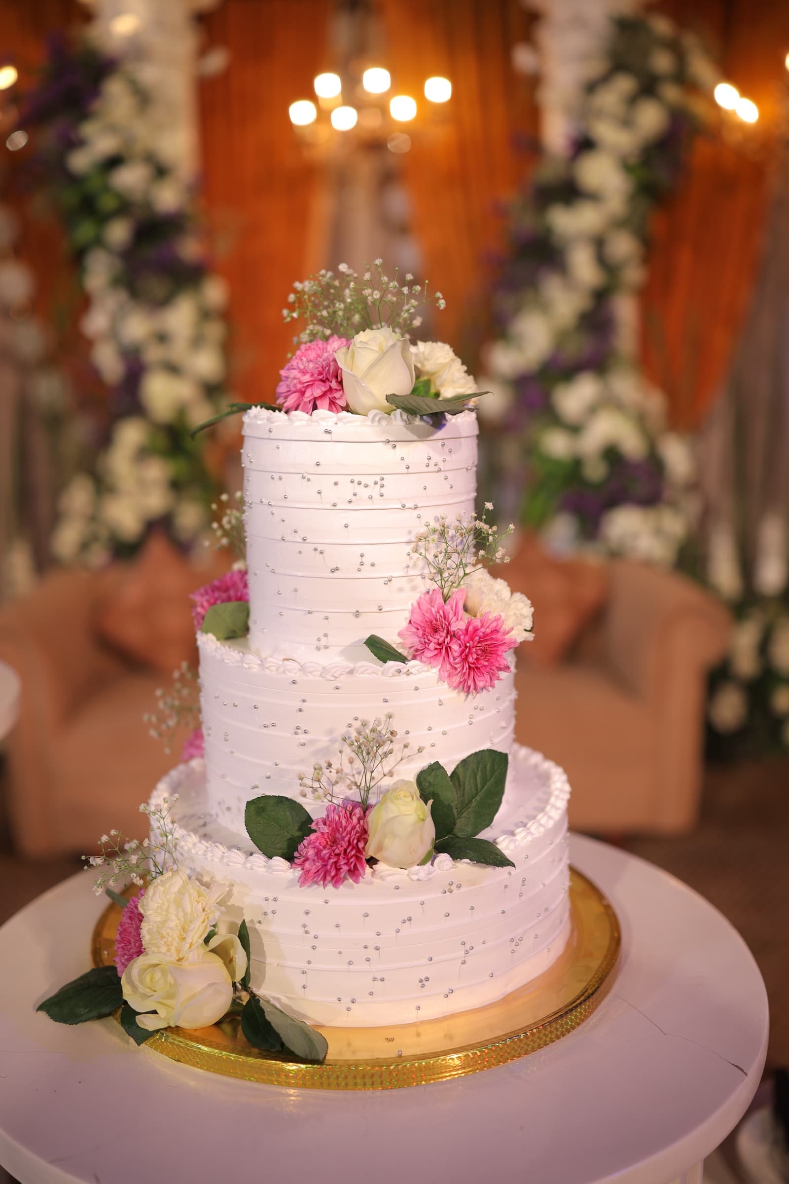 A three-tier white wedding cake with silver pearls, pink dahlias, and white roses