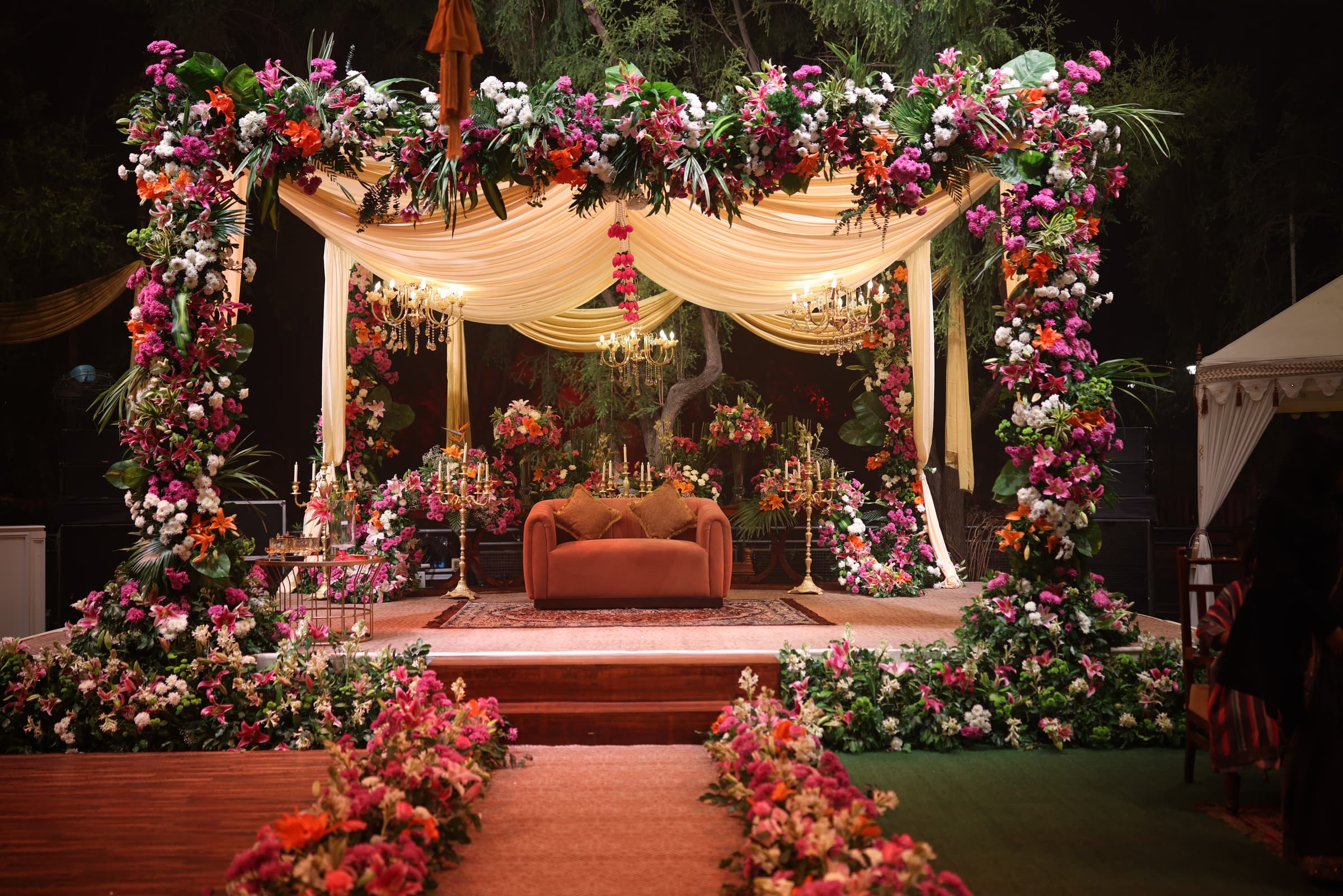 The floral mandap draped in blush and white blooms, with candelabras and a velvet loveseat at the altar