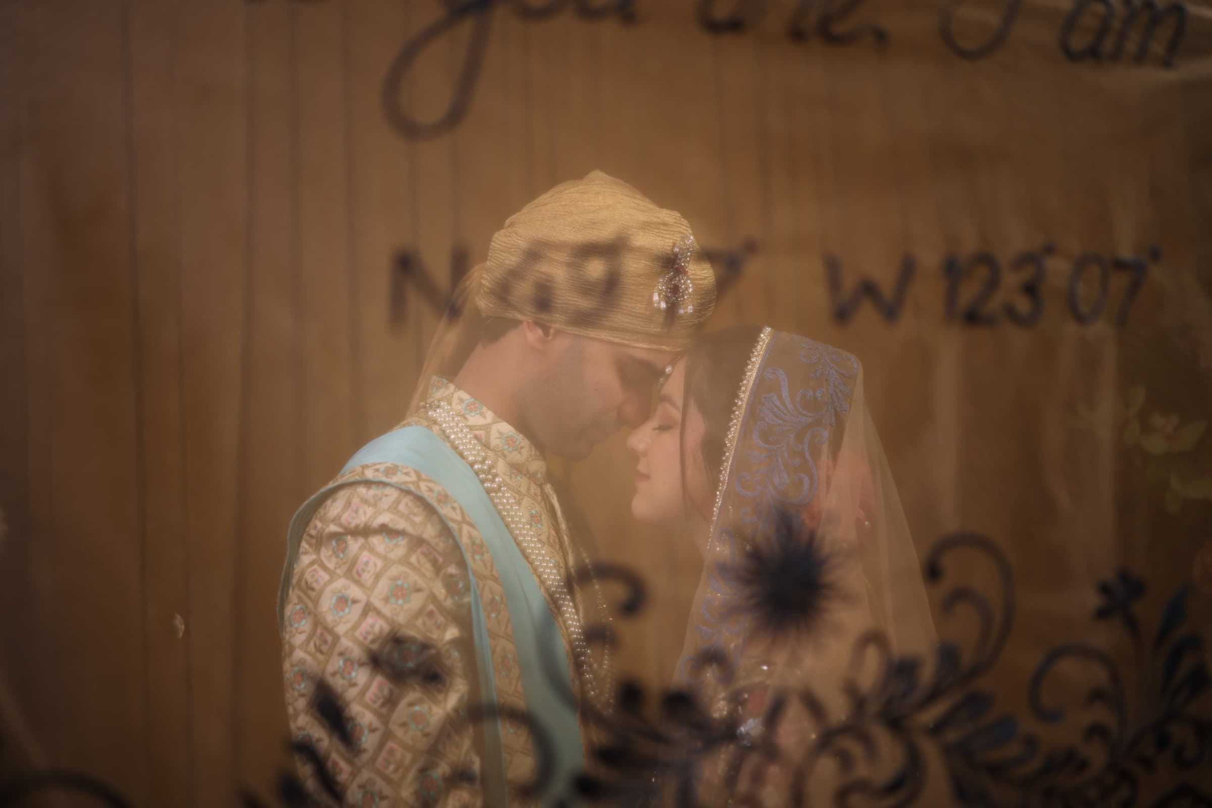 The couple foreheads-to-forehead behind a sheer veil, reflected over hand-lettered venue coordinates