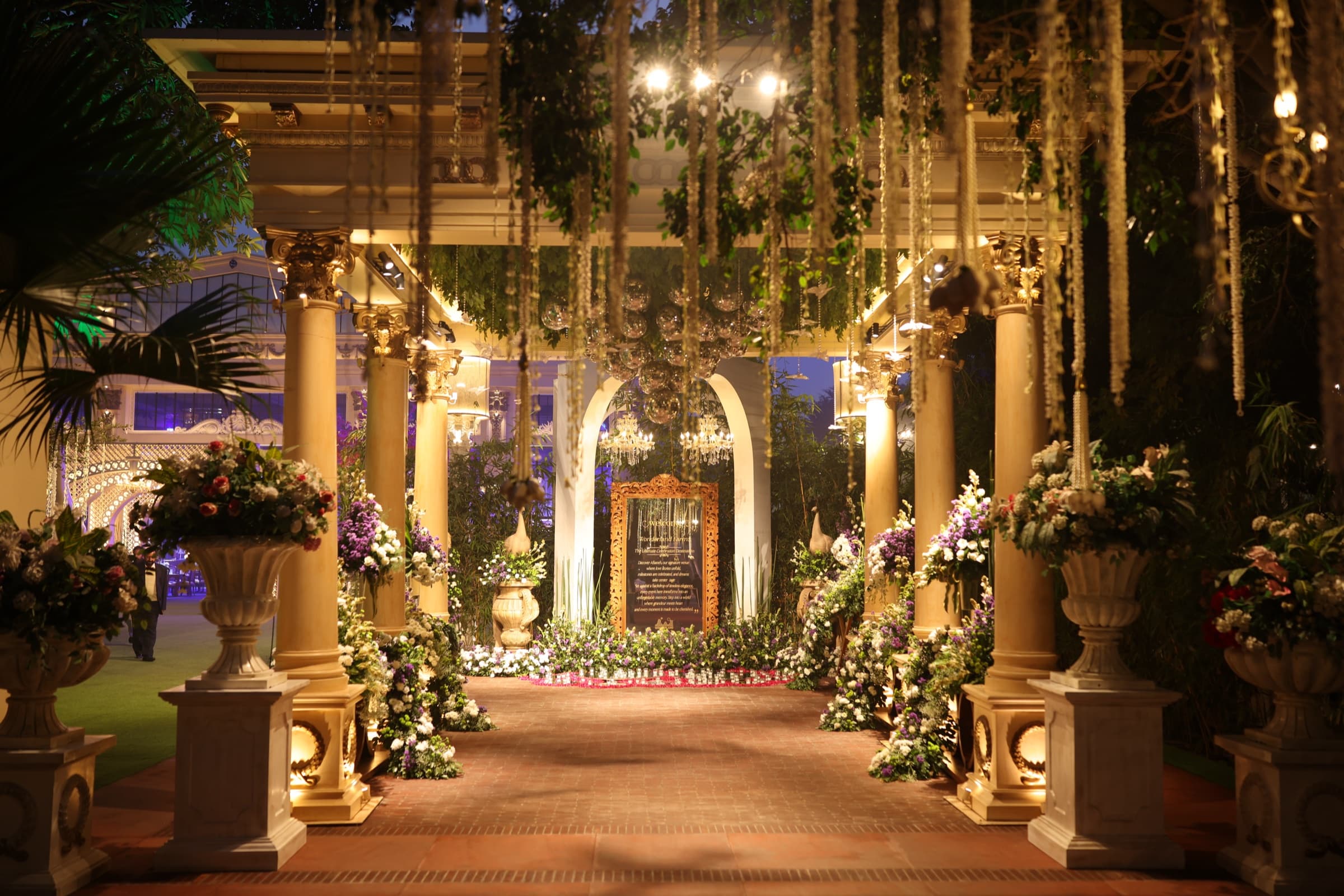 A glowing colonnade entrance with columns, hanging greenery, chandeliers, and a floral-lined pathway