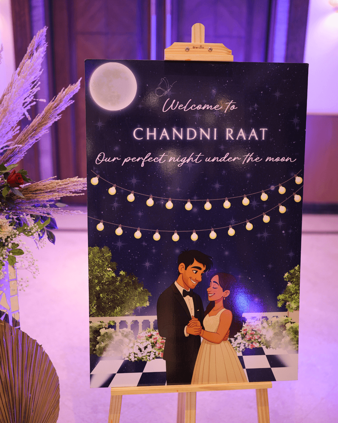 The Chandni Raat cocktail welcome board on an easel under purple venue lighting